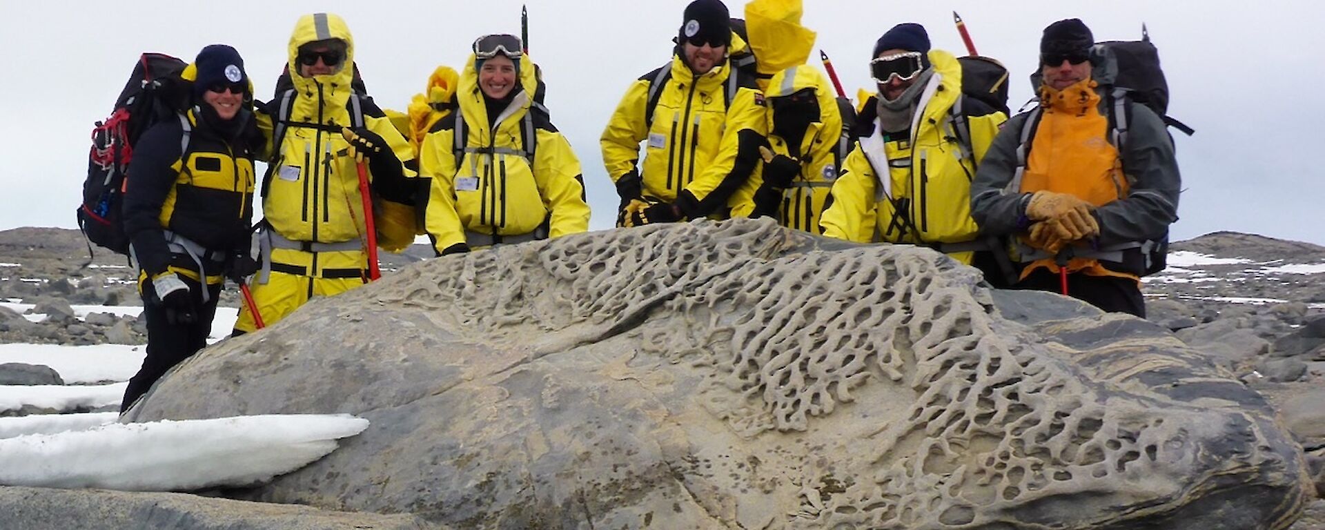 A group in the field standing behind a big rock.
