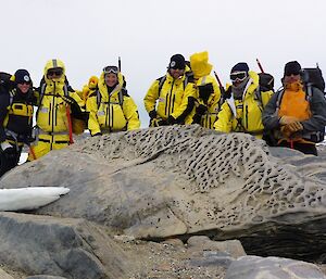 A group in the field standing behind a big rock.