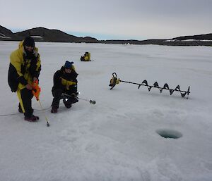 Two people, one kneeling, on sea ice with a drill in the background and round hole in the foreground.