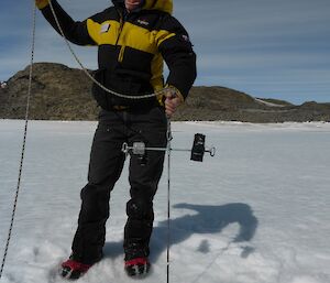 Lady dressed in yellow and black puts a rope down through a hold in the sea ice.