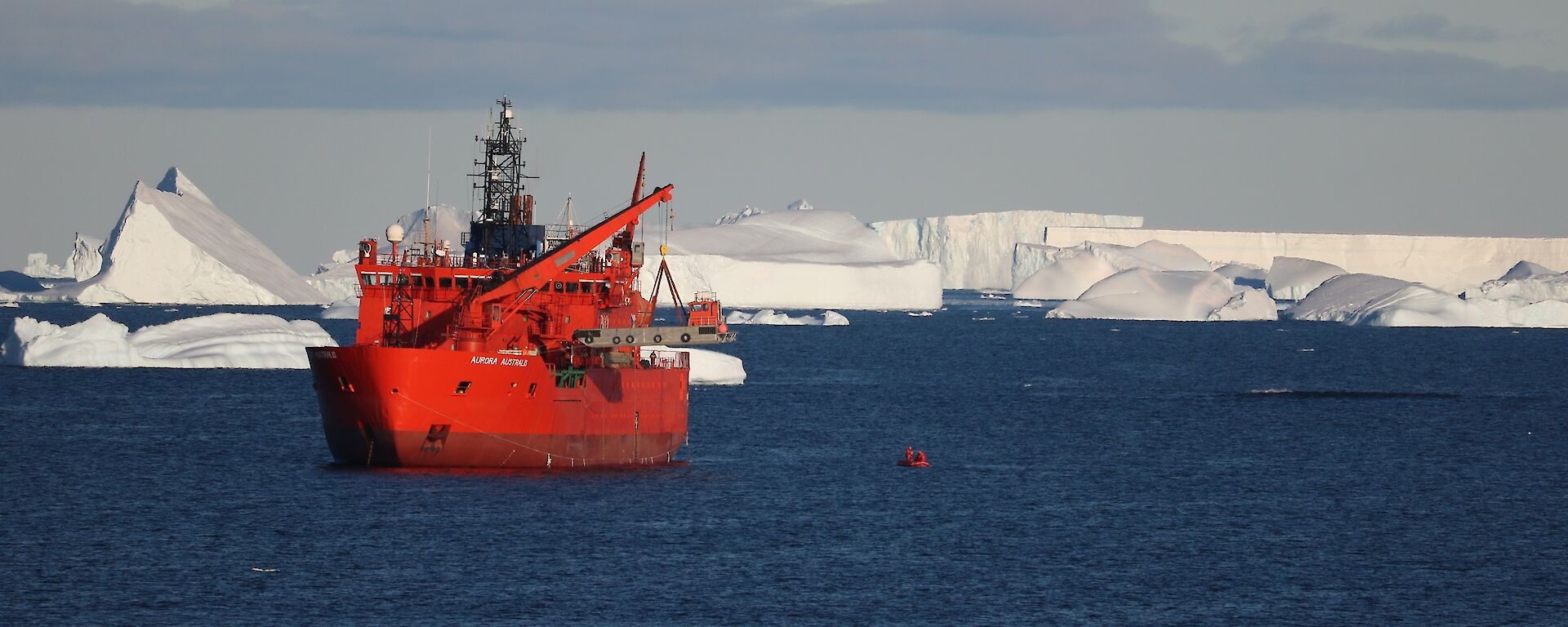 The bright colored Aurora Australis lifting a large jet barge aboard with a small red inflatable rubber boat on the water to the right and large ice burgs in the background.