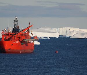 The bright colored Aurora Australis lifting a large jet barge aboard with a small red inflatable rubber boat on the water to the right and large ice burgs in the background.