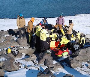 The team gather around the acting patient with a back drop of Heidemann Bays blue water and across the bay ribbons of dolerite dykes can be seen on the rocky hills.