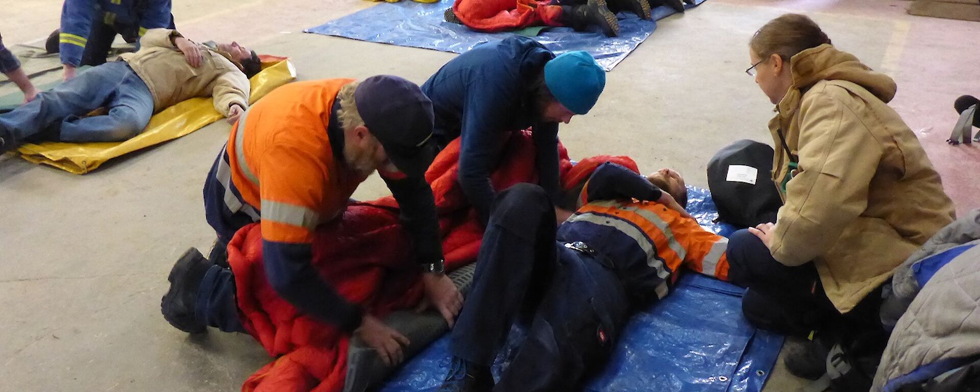 Expeditioners in groups of 3 with a patient who is acting out an injury is rolled into a sleeping bag and onto a blue matt.