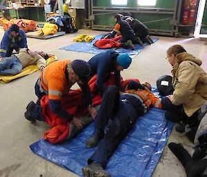 Expeditioners in groups of 3 with a patient who is acting out an injury is rolled into a sleeping bag and onto a blue matt.