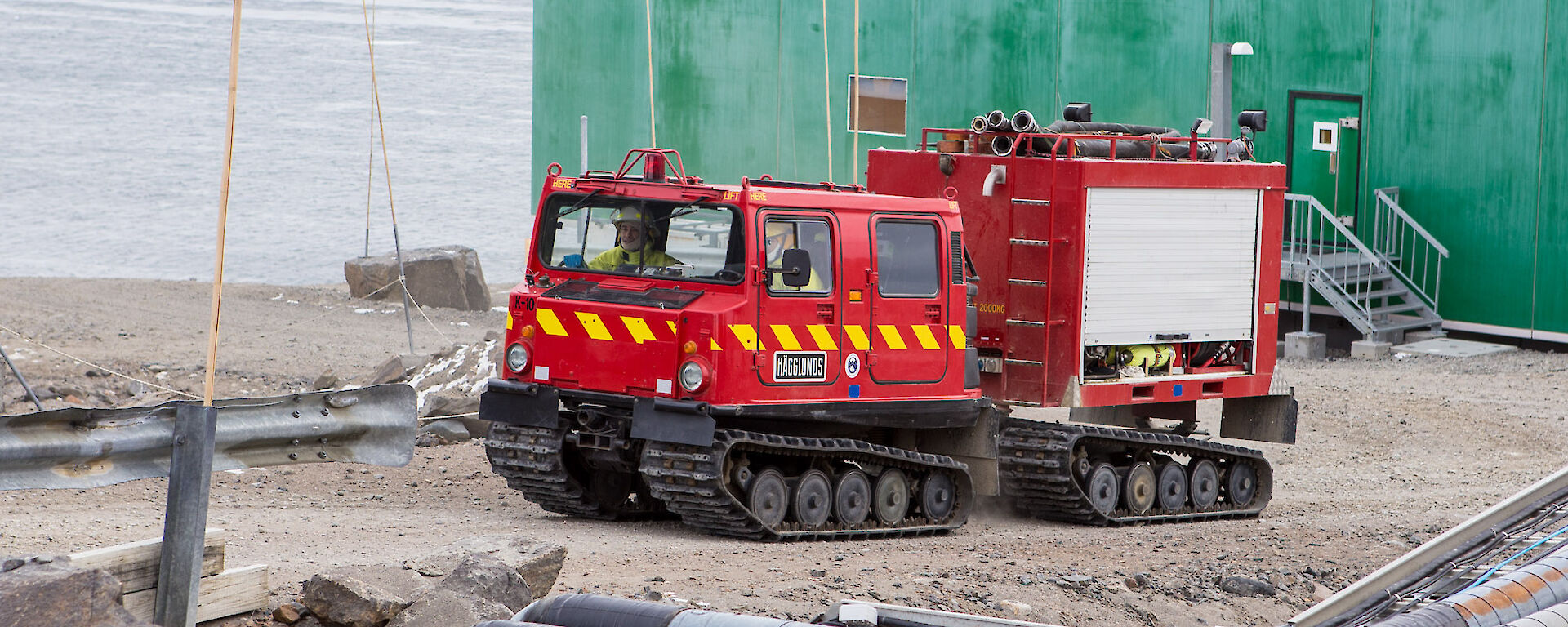 The bright red fire hagglunds vehicle driving up the hill towards the summer accommodation building.