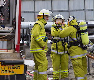Two fire team members dressed in full firefighting turnout protective clothing donning breathing apparatus.