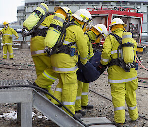 Four fire team members carry the dummy who is dressed in blue overalls out of the building.