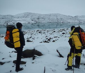 Two people with back packs looking at snow covered hills and lake with a reflection image of the hill.