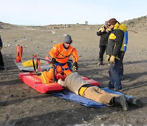 One expeditioner in a bright orange dry suit practicing to manoeuvre a casualty onto the recue alive unit while still on shore.