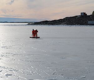 Two expeditioner in bright orange dry suits drilling the ice 60 metres from shore with some station buildings in the background.