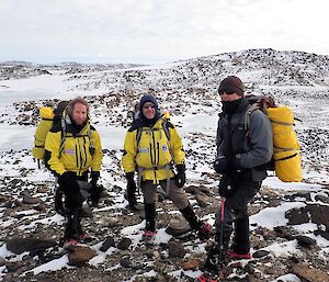 Three expeditioners posing for a photo with their back packs and wind protective clothing and the rocky Vestfold hills in the background.