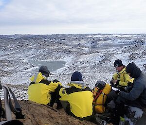 Four expeditioners all sitting together have a drink with the rolling snow covered rocky Vestfold Hills in the background.