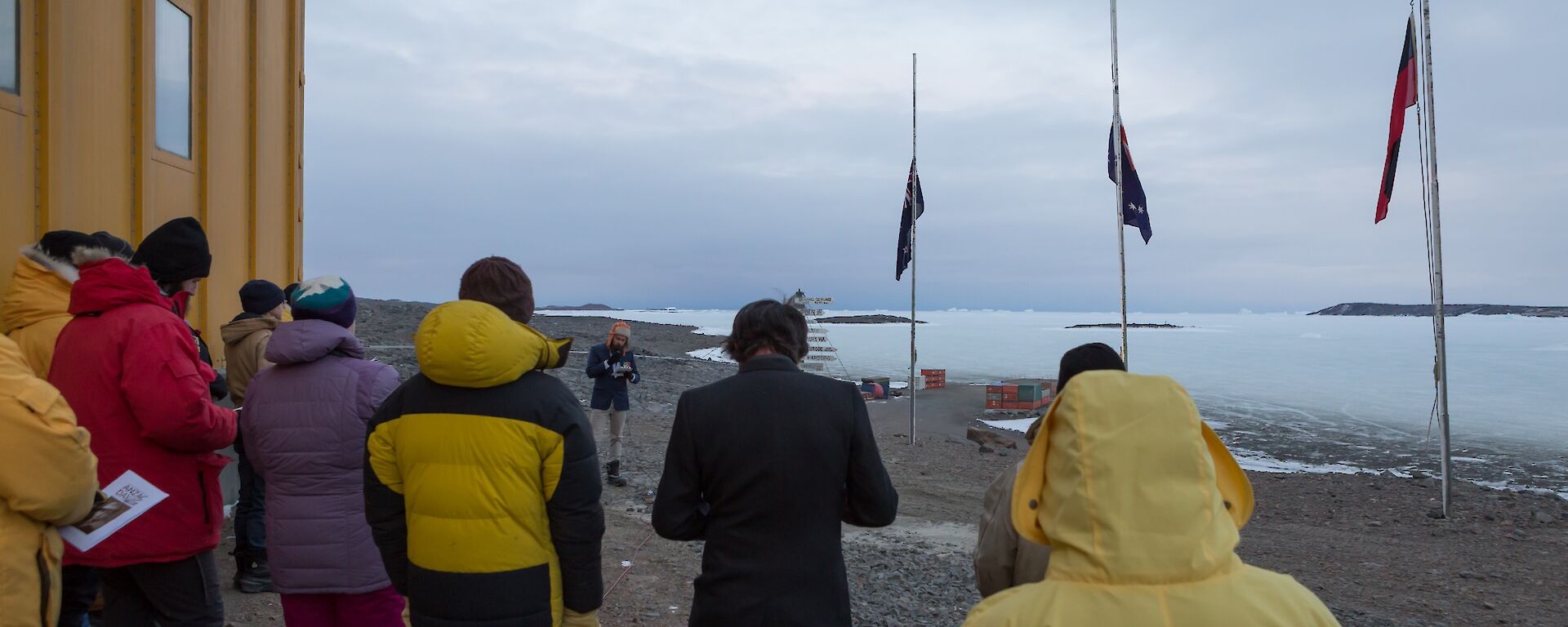 Expeditioners conducting the Anzac dawn service at the station flagpoles with the Australian, aboriginal and New Zealand flags at half-mast.