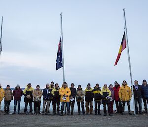 A group photo of the Davis team at the station flagpoles with the Australian, Aboriginal and New Zealand flags at half-mast.