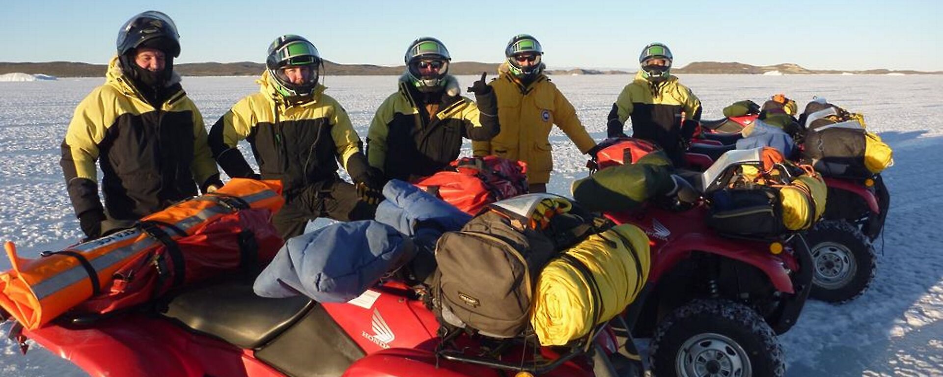 A Line up of Quad bikes with expeditioners standing behind them on the sea ice under clear blue skies.