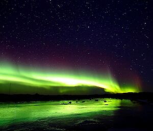 Bright green aurora reflecting off a frozen freshwater lake with a star filled sky background.