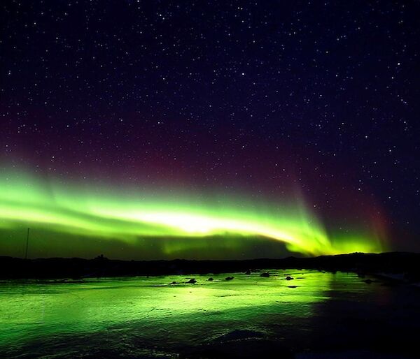 Bright green aurora reflecting off a frozen freshwater lake with a star filled sky background.