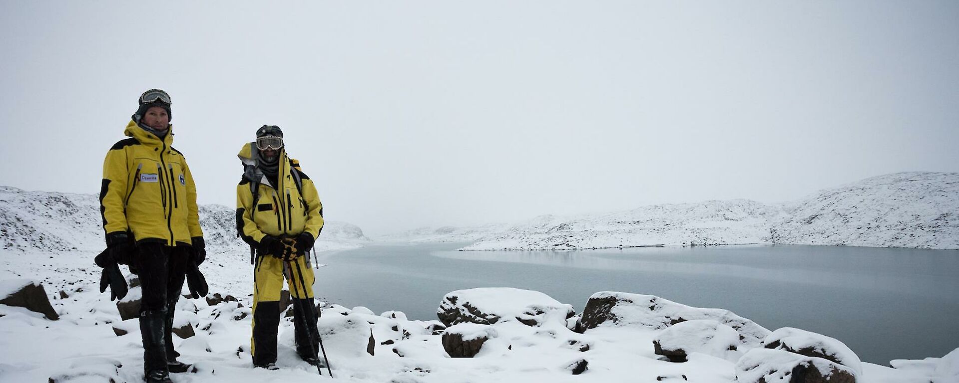 Two expeditioners dressed in yellow wind proof clothing with the snow covered hills and Deep Lake in the background.