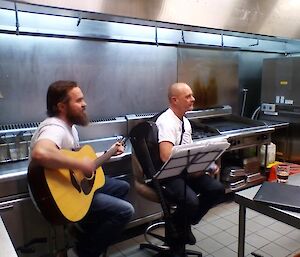 Chris and Rodney sitting in front of the kitchen equipment playing their guitars.