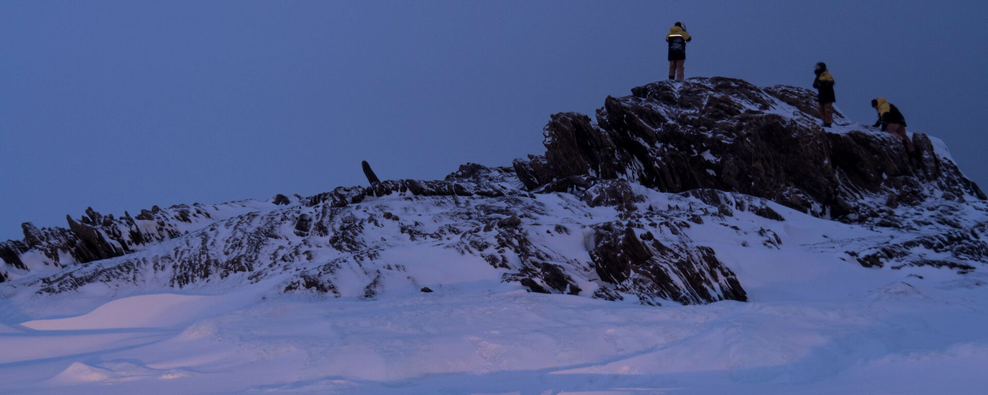 Three expeditioners standing on top a small rocky hill which is surrounded by sea ice and snow.