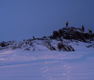 Three expeditioners standing on top a small rocky hill which is surrounded by sea ice and snow.