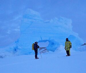 Two expeditioners on cross country skis in front of a small ice berg with a large hole in the middle.
