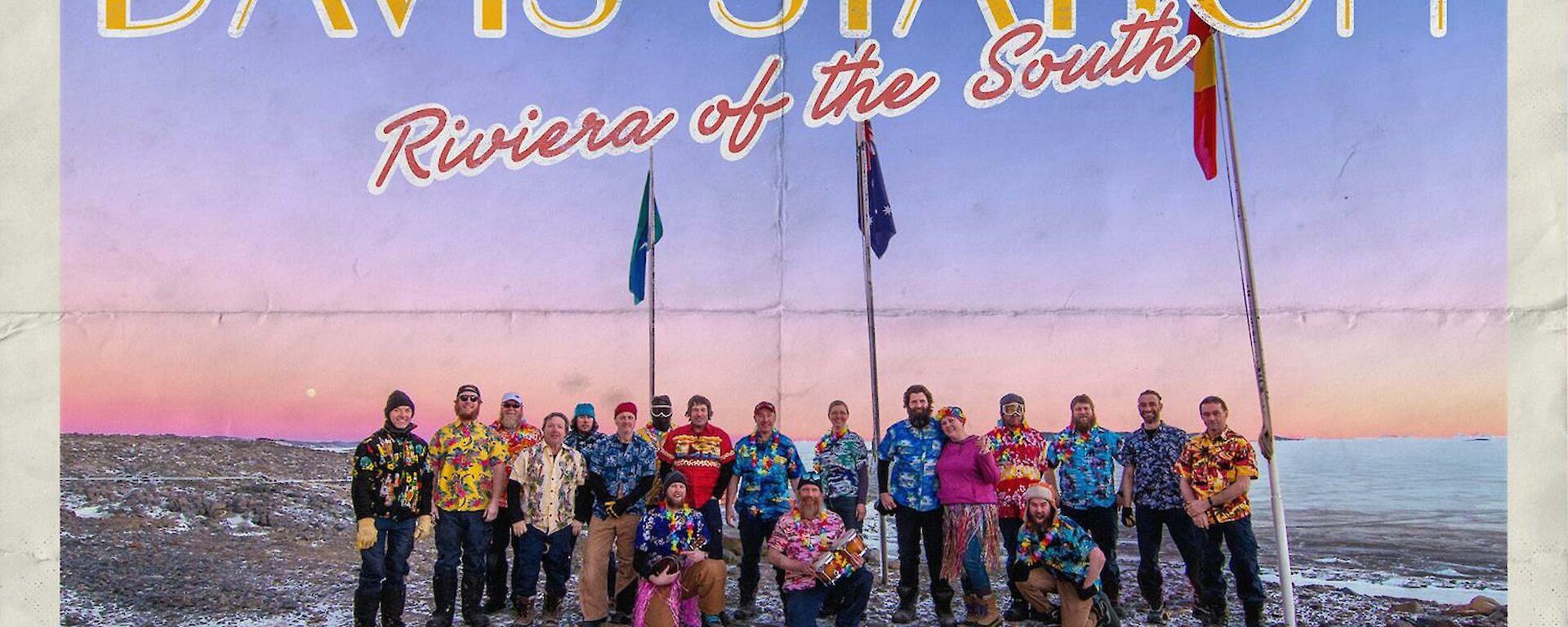 Group photo of station expeditioners all wearing Hawaiian shirts under the flag poles on a blue sky day with a red sun haze on the horizon.