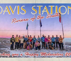 Group photo of station expeditioners all wearing Hawaiian shirts under the flag poles on a blue sky day with a red sun haze on the horizon.
