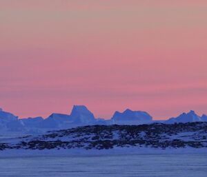The pink hues of sunset colour the sky behind the islands and icebergs.