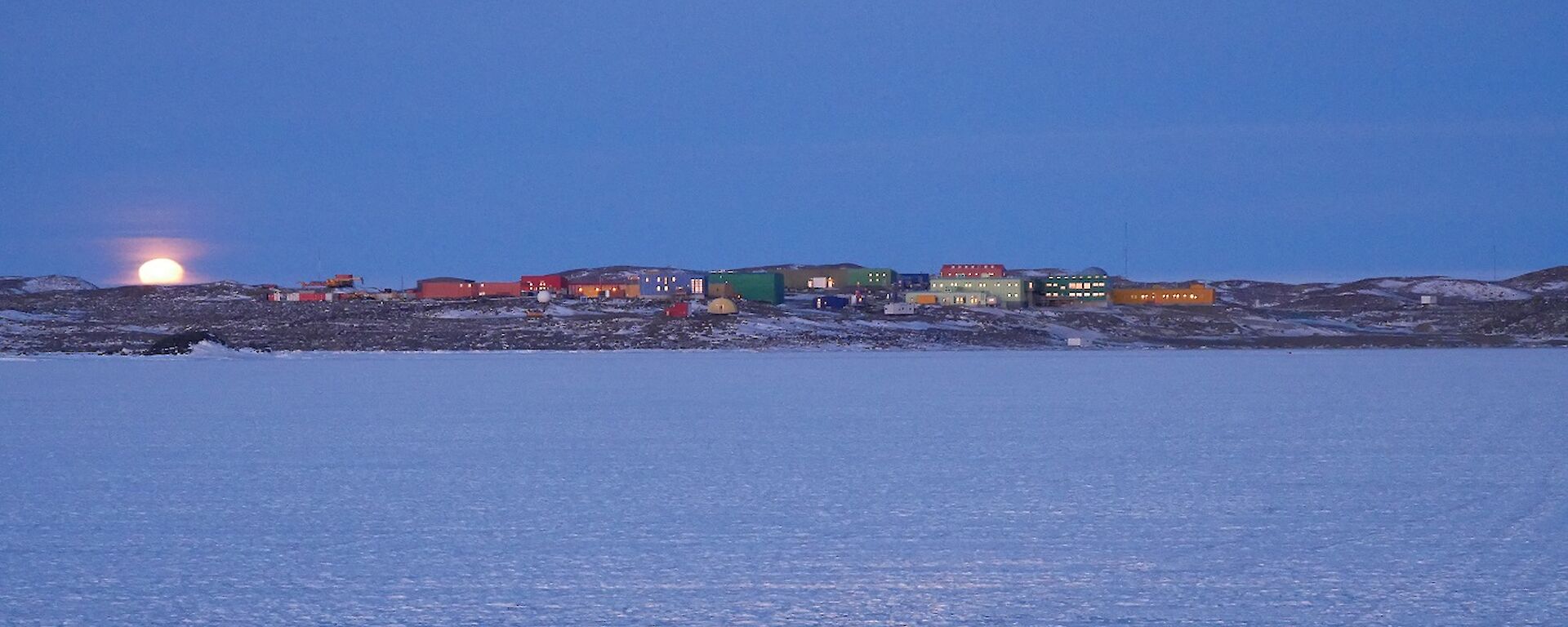 Coloured buildings in snow and rock landscape with moon rising over hills in background