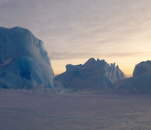 Three majestic icebergs with a glow in the sky behind them. The left berg consists of clear crystal blue ice.