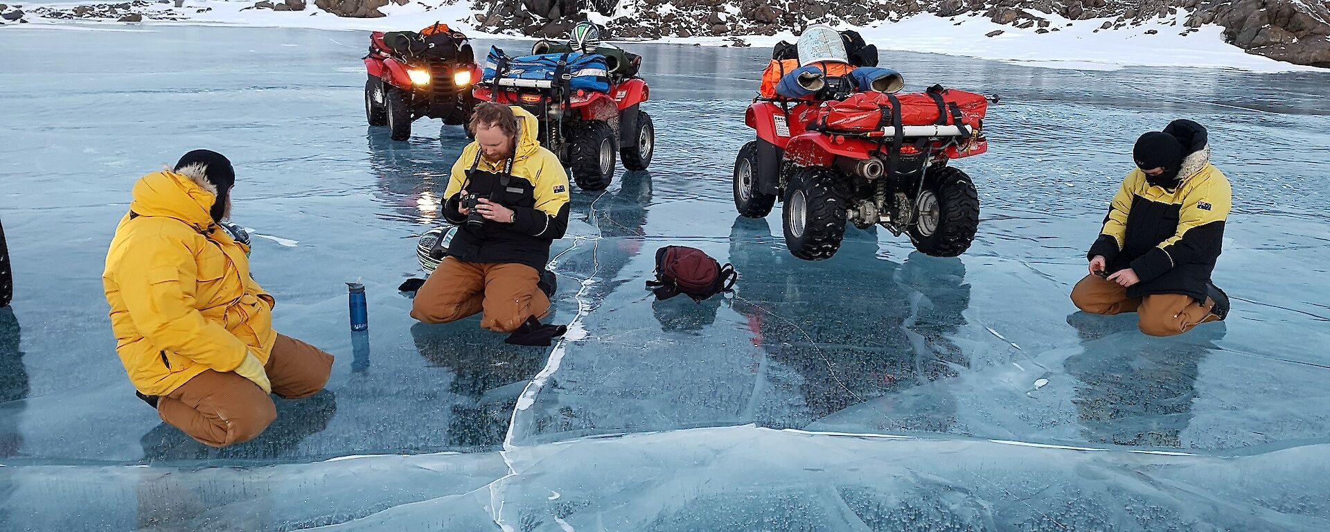 Three expeditioners of their quad bikes and on their knees taking photos of the air bubbles trapped in the frozen lake.