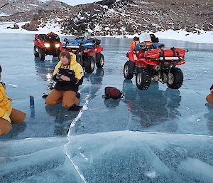 Three expeditioners of their quad bikes and on their knees taking photos of the air bubbles trapped in the frozen lake.