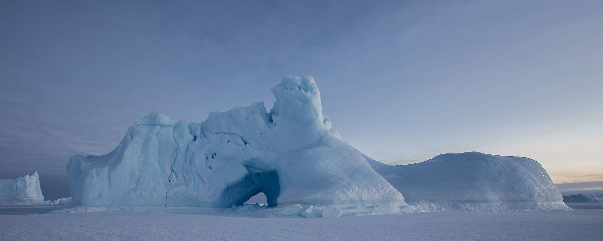 An ice burg locked in the sea ice that has a large hole weathered in the side.