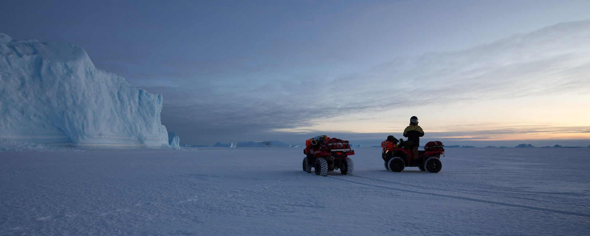 Two quad bikes on the snow covered sea ice, with large ice bergs in the background.