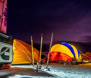 Bright coloured tents pitched on the snow in the late evening with lights on.