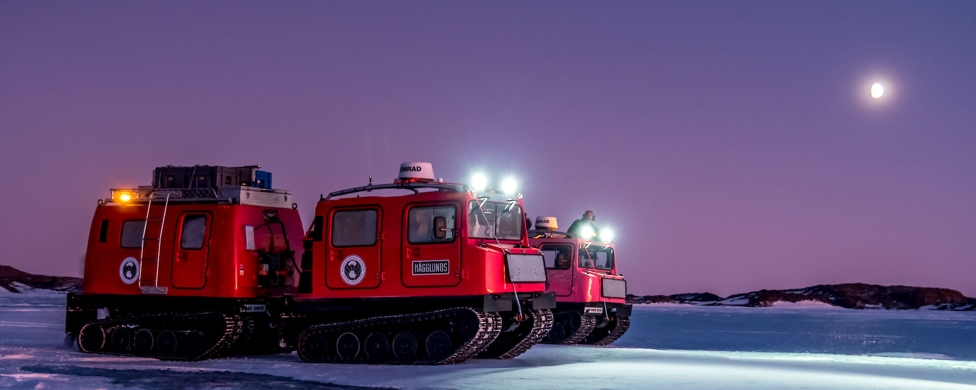 Red and pink Hägglunds vehicles on the sea ice with headlights on and the moon in the distant sky.