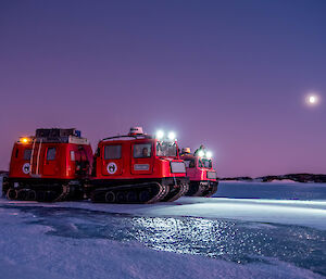 Red and pink Hägglunds vehicles on the sea ice with headlights on and the moon in the distant sky.