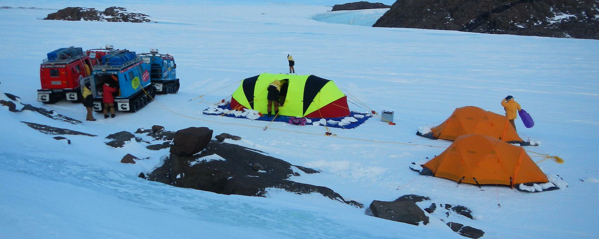 The campground, with the red and blue Hägglunds vehicles parked on the left and fluorescent striped oval shaped Endurance tent is in the middle of the two orange Polar Dome tents on the right.