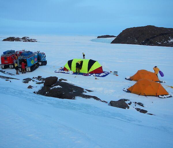 The campground, with the red and blue Hägglunds vehicles parked on the left and fluorescent striped oval shaped Endurance tent is in the middle of the two orange Polar Dome tents on the right.