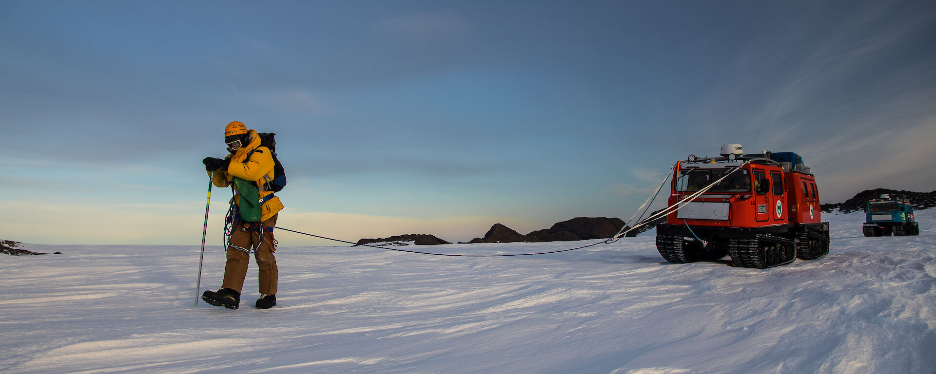 Derryn practicing probing for crevasses wielding a long metal spike while connected by a rope to the red Hägglunds vehicle.