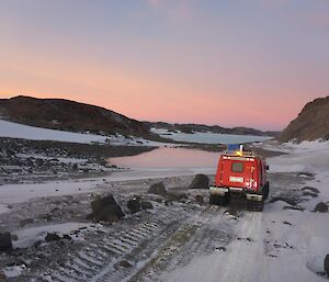 Red Hägglunds vehicle driving across a snow covered dirt track at Pioneer Crossing, a short land bridge between two bays of sea ice. Pink and purple sky at dawn on the horizon.