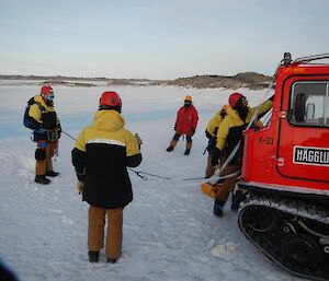 Chris harnessed up and connected to the red Hägglunds vehicle by rope ready to lower himself off the edge of a snowy hill while Simon, Richard, Iain and Aaron helping with preparations in front of the red Hägglunds.