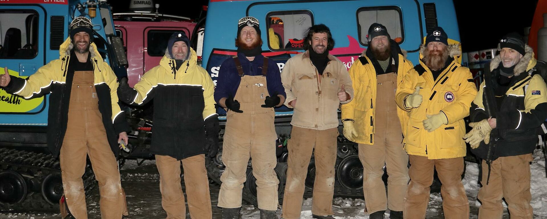 The crew standing along the side of the blue Hagglund vehicle, with big smiles and thumbs up, about to depart in the dark of early morning