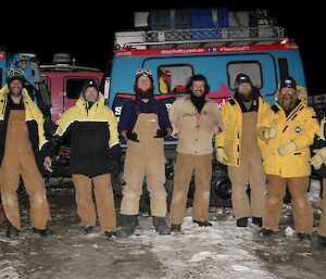 The crew standing along the side of the blue Hagglund vehicle, with big smiles and thumbs up, about to depart in the dark of early morning