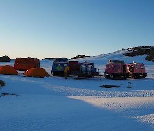 Our campsite on the snow in amongst some low rocky hills in the evening light, orange RMIT van and blue Hägglunds vehicle at the back, two orange polar dome tents in front and the pink Hägglund vehicle to the right