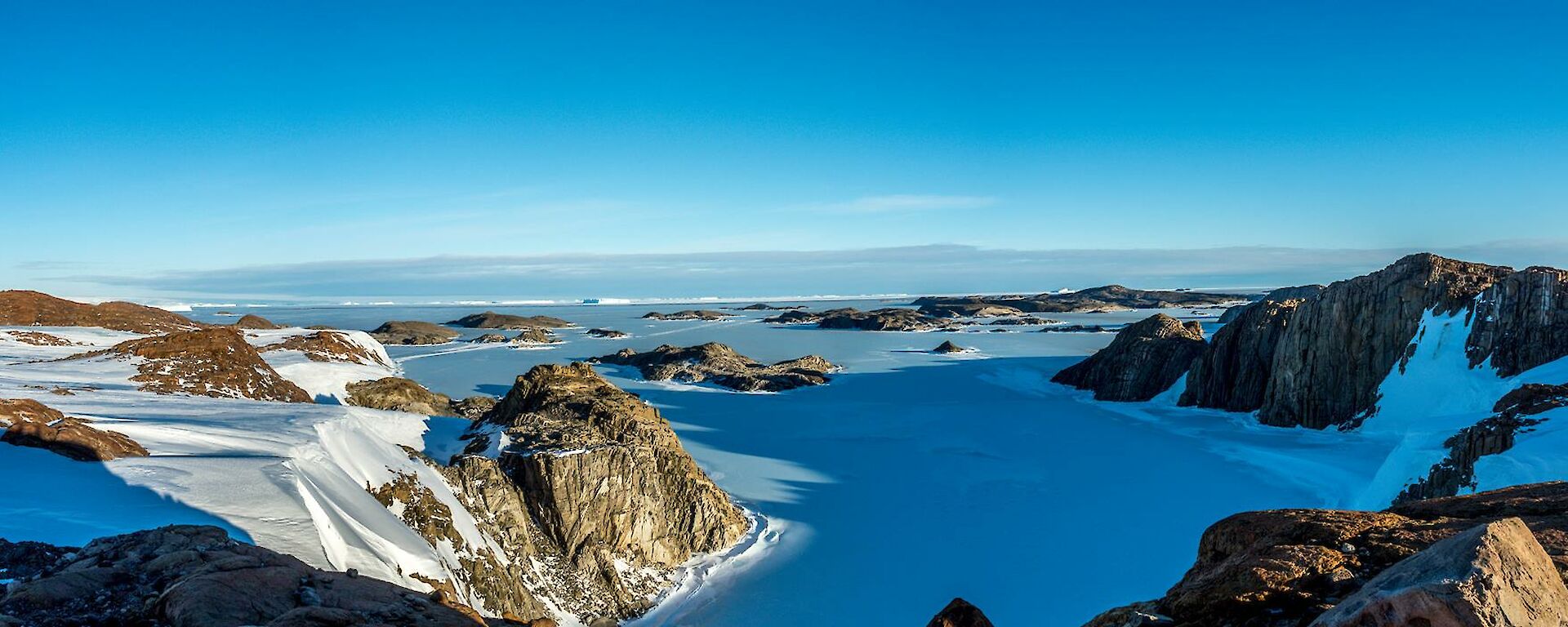 Panoramic view across the Rauer Islands from an elevated position. The hills and islands are in sunlight and the hills are casting long shadows across the sea ice between the islands.