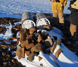 Picture of a cache from 2002, with two fuel drums and a pile of rocks weighing down a tent and a container of food.