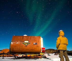 Starry night sky with a ribbon of green aurora over the small orange caravan-like RMIT van with Dan is standing in the bottom right wearing a yellow down hooded jacket
